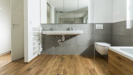 Ein modernes Badezimmer mit einem Holzfußboden. An der linken Wand befindet sich ein Handtuchheizkörper. Die Wand hinter dem Waschbecken ist mit großen Spiegeln verkleidet. Rechts im Bild sind eine Toilette und ein Teil einer Badewanne zu sehen. Die Wände sind in einem schlichten, grauen Farbton gehalten.