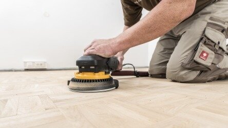 Ein Handwerker kniet auf einem Holzboden und benutzt eine elektrische Schleifmaschine, um den Boden zu bearbeiten. Der Boden hat ein Fischgrätmuster, und im Hintergrund ist eine weiße Wand mit einer Steckdose zu sehen. Der Handwerker trägt beige Arbeitskleidung.