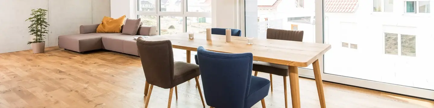 Ein modernes, helles Wohnzimmer mit Essbereich und großen Fenstern. Im Vordergrund steht ein Holztisch mit vier unterschiedlich farbigen Stühlen. Im Hintergrund befindet sich ein graues Ecksofa mit gelbem Kissen und eine grüne Zimmerpflanze. Der Raum wirkt freundlich und minimalistisch eingerichtet.