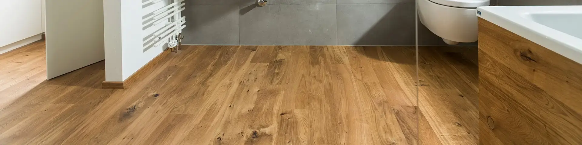Parkettboden in einem Badezimmer. Es sind Teile der Badewanne, Toilette und einer Heizung zu sehen.