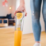 Nahaufnahme des Spray Mop Flüssigkeitsbehälters. Dieser wurde von einer Person an der richtigen Stelle des Wischmopps eingesetzt. Im Hintergrund ist das moderne Wohnzimmer zu sehen.
